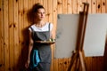 Woman with paints stands on wooden wall background and blurred easel in foreground. Royalty Free Stock Photo