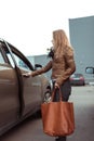 Woman in parking lot of shopping center opens car door, in her hand is bag with groceries and purchases, brown leather