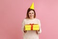 Woman in party cone holding yellow wrapped gift box, looking at camera with satisfied expression. Royalty Free Stock Photo