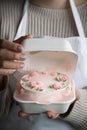 Woman pastry shef wearing white apron and holding small bento cake decorated with cream cheese pink flowers. Woman hands hold Royalty Free Stock Photo