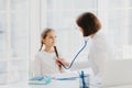 Woman pediatrician gives consultation to small girl, talk about health and symptoms, listens heart with phonendoscope, makes