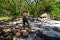 A woman photographs a water landscape using a tablet, standing on stones among a small river.