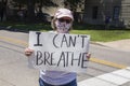 Woman in pink ball cap and printed mask holding sign reading I cant breathe standing in middle of road