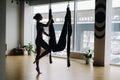 A woman in pink clothes does yoga on a suspended burgundy hammock in a bright gym