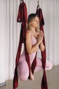A woman in pink clothes does yoga on a suspended burgundy hammock in a bright gym