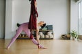 A woman in pink clothes does yoga on a suspended burgundy hammock in a bright gym