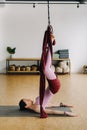 A woman in pink clothes does yoga on a suspended burgundy hammock in a bright gym