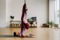 A woman in pink clothes does yoga on a suspended burgundy hammock in a bright gym