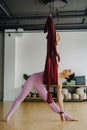 A woman in pink clothes does yoga on a suspended burgundy hammock in a bright gym