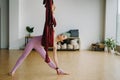 A woman in pink clothes does yoga on a suspended burgundy hammock in a bright gym