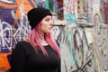 Woman with pink hair piercings and tattoos leaning against graffiti wall