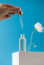 A woman with a pipette drips skin care serum into a transparent dropper, which stands on a podium on a blue background