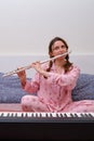 A woman plays a flute sitting on a home bed at an electric piano