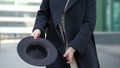 Woman plays with modern black hat moving wide-brimmed chapeau at black coat bottom in street slow motion low angle shot.