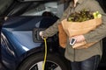 Woman plugs a charger to electric car socket at home after shopping groceries.