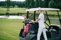 woman in polo and cap with golf gear standing at golf cart at golf course