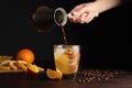 Woman pouring coffee into glass with orange juice and ice cubes at wooden table, closeup Royalty Free Stock Photo