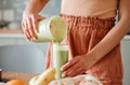 Woman pouring healthy smoothie in a glass from a blender jar on a counter for detox. Female making fresh green fruit