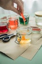 Woman pouring liquid orange Easter cold colour color in glass with water and vinegar next to green, yellow, red, blue dye in Royalty Free Stock Photo