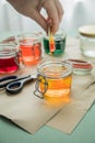 Woman pouring liquid orange Easter cold colour color in glass with water and vinegar next to green, yellow, red, blue dye in Royalty Free Stock Photo