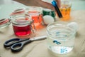 Woman pouring liquid orange Easter cold colour color in glass with water and vinegar next to green, yellow, red, blue dye in Royalty Free Stock Photo