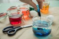 Woman pouring liquid orange Easter cold colour color in glass with water and vinegar next to green, yellow, red, blue dye in Royalty Free Stock Photo