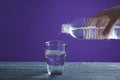 Woman pouring water from bottle into glass on wooden table against purple background, closeup Royalty Free Stock Photo