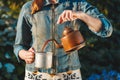 A woman pours delicious coffee from an antique kettle into an aluminum mug. The background is blurred