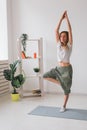 Woman practise yoga performs. Pose on mat inside of cozy room with plants and greenery. Healthy lifestyle concept.