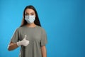 Woman in protective face mask and medical gloves showing thumb up gesture on blue background. Space for text Royalty Free Stock Photo