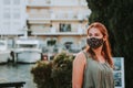 Woman in protective fashionable face mask with smartphone in a touristic place during covid new normal with crowded street cafe in