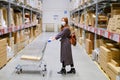 A woman in a protective mask with a cart in a market. Redhead girl in a store warehouse during a coronavirus epidemic, concept