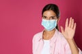 Woman in protective mask showing hello gesture on pink background, space for text. Keeping social distance during coronavirus