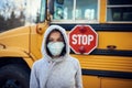 A woman in a protective mask stands on the background of a school bus. A large stop sign is visible on the background