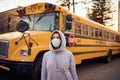 A woman in a protective mask stands on the background of a school bus. A large stop sign is visible on the background