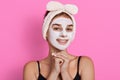 Woman with purifying white mask on her face, cute female with charming smile keeping hands under chin, beauty procedures at home