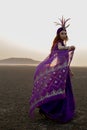 a woman in a purple dress and feathered hat is standing in the desert Royalty Free Stock Photo