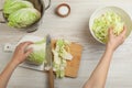 Woman putting cut Chinese cabbage into bowl at white wooden kitchen table, top view