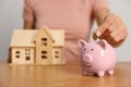 Woman putting money into piggy bank and holding house model at wooden table, focus on hand Royalty Free Stock Photo