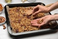 Woman putting nuts into tray with tasty granola at white marble table, closeup