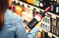 Woman reading the label of red wine bottle in liquor store or alcohol section of supermarket. Shelf full of alcoholic beverages. Royalty Free Stock Photo