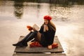 Woman in a red beret reading book on wooden pontoon. Autumn season. Royalty Free Stock Photo