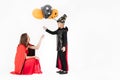 Woman in red costume giving fancy balloons to kid boy in back suit on white background. Concept for funny playing in halloween fe