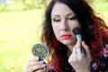 Woman with red curly hair corrects her make-up on the green lawn. puts the powder with a brush looking into a mirror Royalty Free Stock Photo