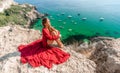 Woman red dress sea. Happy woman in a red dress and white bikini sitting on a rocky outcrop, gazing out at the sea with Royalty Free Stock Photo