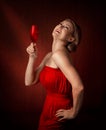 A woman in a red dress stands on a brown background and holds a wine glass in her hand. Middle aged blonde.