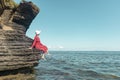 A woman in a red dress and white hat sits on a cliff of a cliff by the sea and looks into the distance at the horizon. Clear sky. Royalty Free Stock Photo