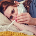 A woman with red eyes, sick with the flu, looks sadly at the thermometer. The patient lies on a bed in a medical mask