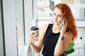 Woman with red hair is drinking coffee and talking on the phone at the workplace in the office Royalty Free Stock Photo