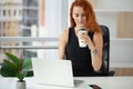 Woman with red hair is drinking coffee and talking on the phone at the workplace in the office Royalty Free Stock Photo